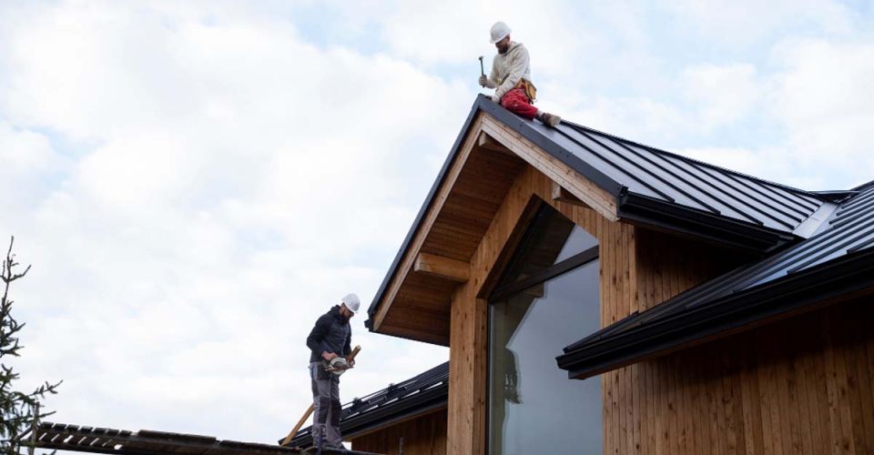 Two men are working together on the roof of a house, focused on their tasks with tools in hand.